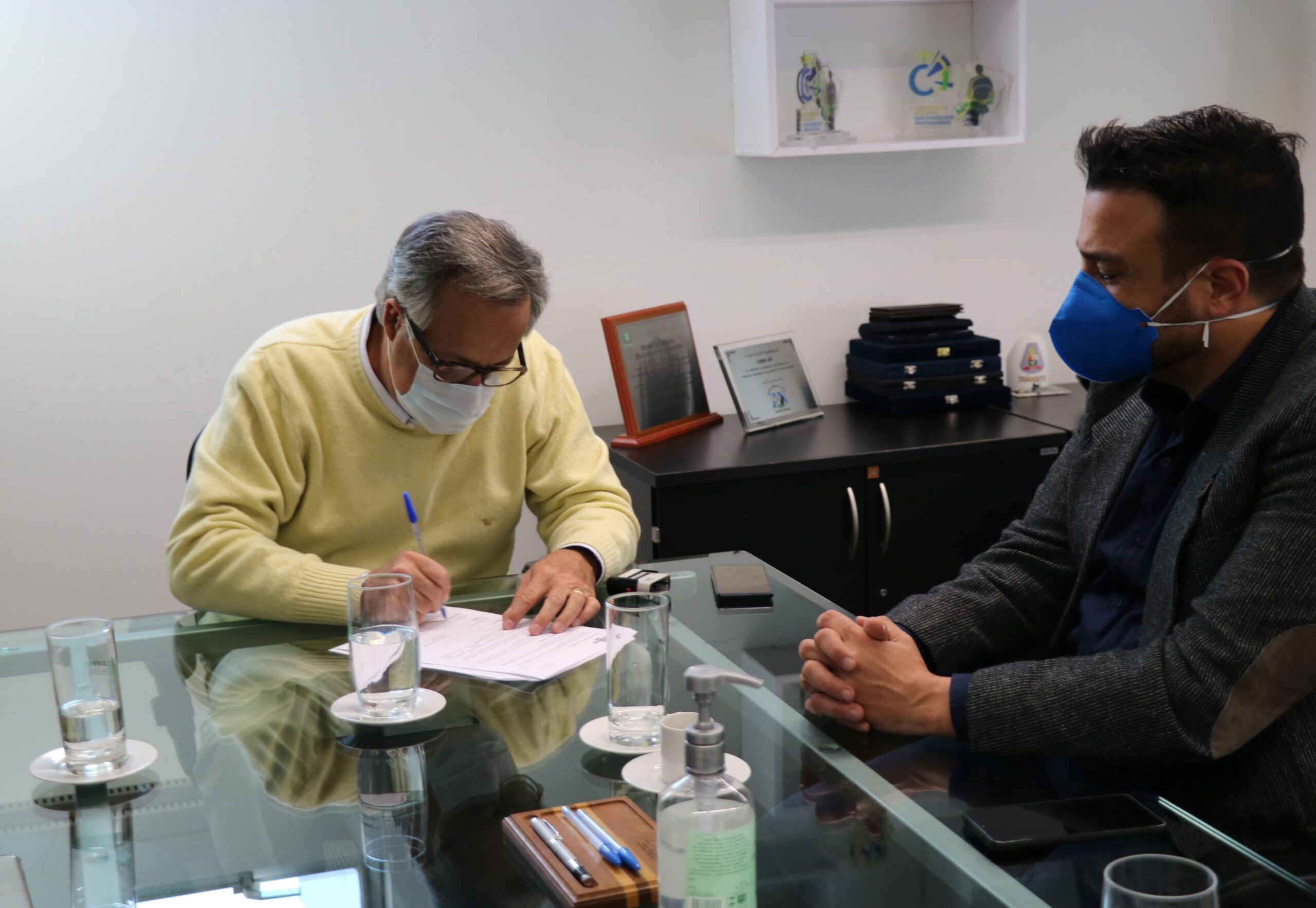 Presidente do CRMV-SP está de suéter bege, máscara e óculos, sentado à mesa assinando o protocolo de intenção, ao lado do gerente regional Osasco do Sebrae.