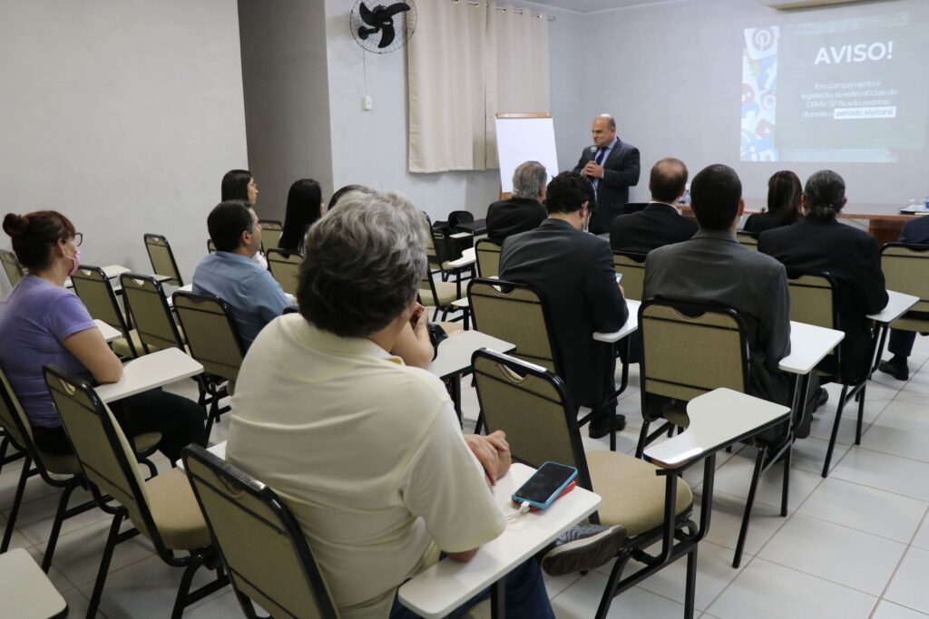 Foto mostra um auditório com carteiras escolares e pessoas nelas sentadas. A frente está o vice-presidente do CRMV-SP falando ao microfone. Ao fundo um telão com aviso sobre as restrições inerentes ao período eleitoral.