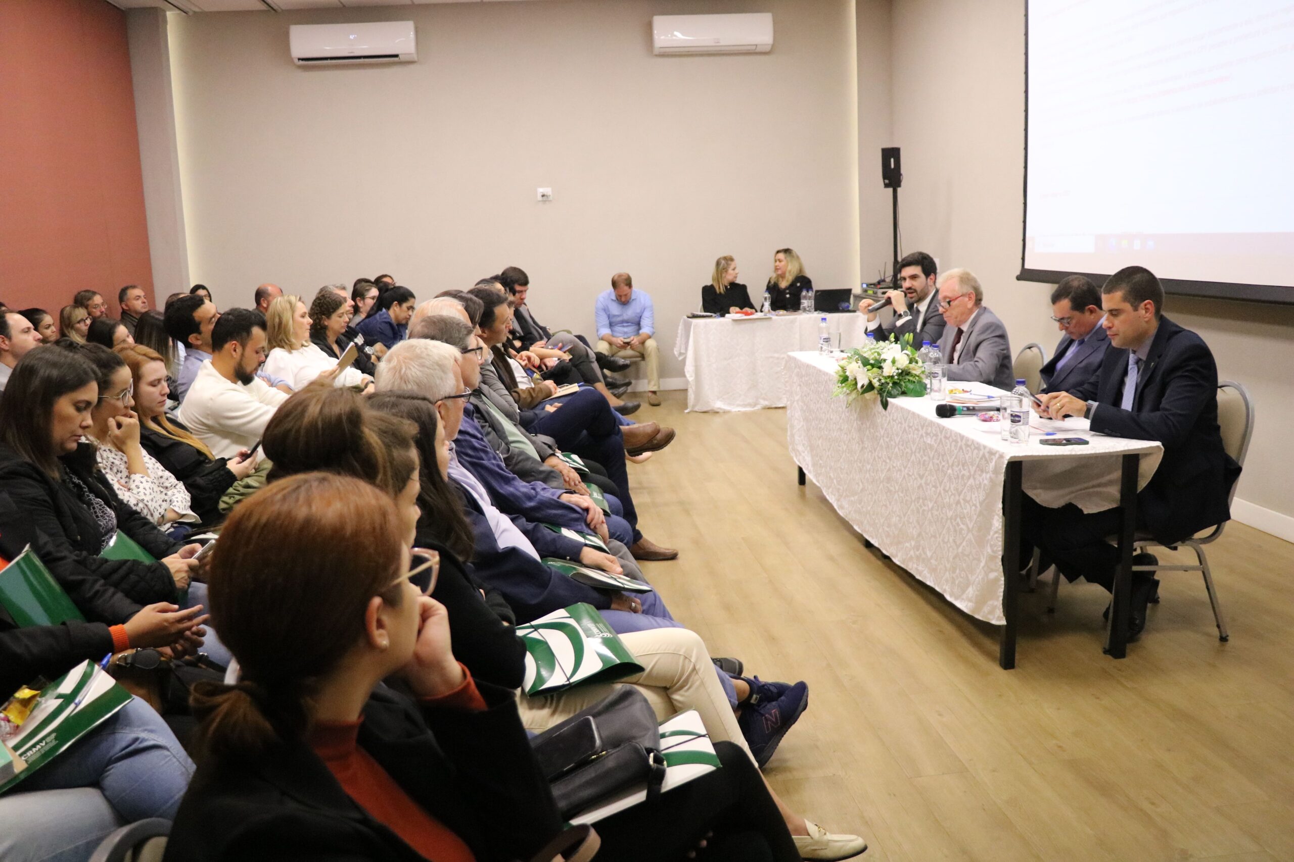 Na imagem aparece o auditório lotado durante o CRMV-SP Escuta de Mogi Mirim. Aparece também o assessor jurídico, o presidente, o secretário-geral e o diretor técnico sentados atrás de uma mesa com toalha branca e arranjo de flores.