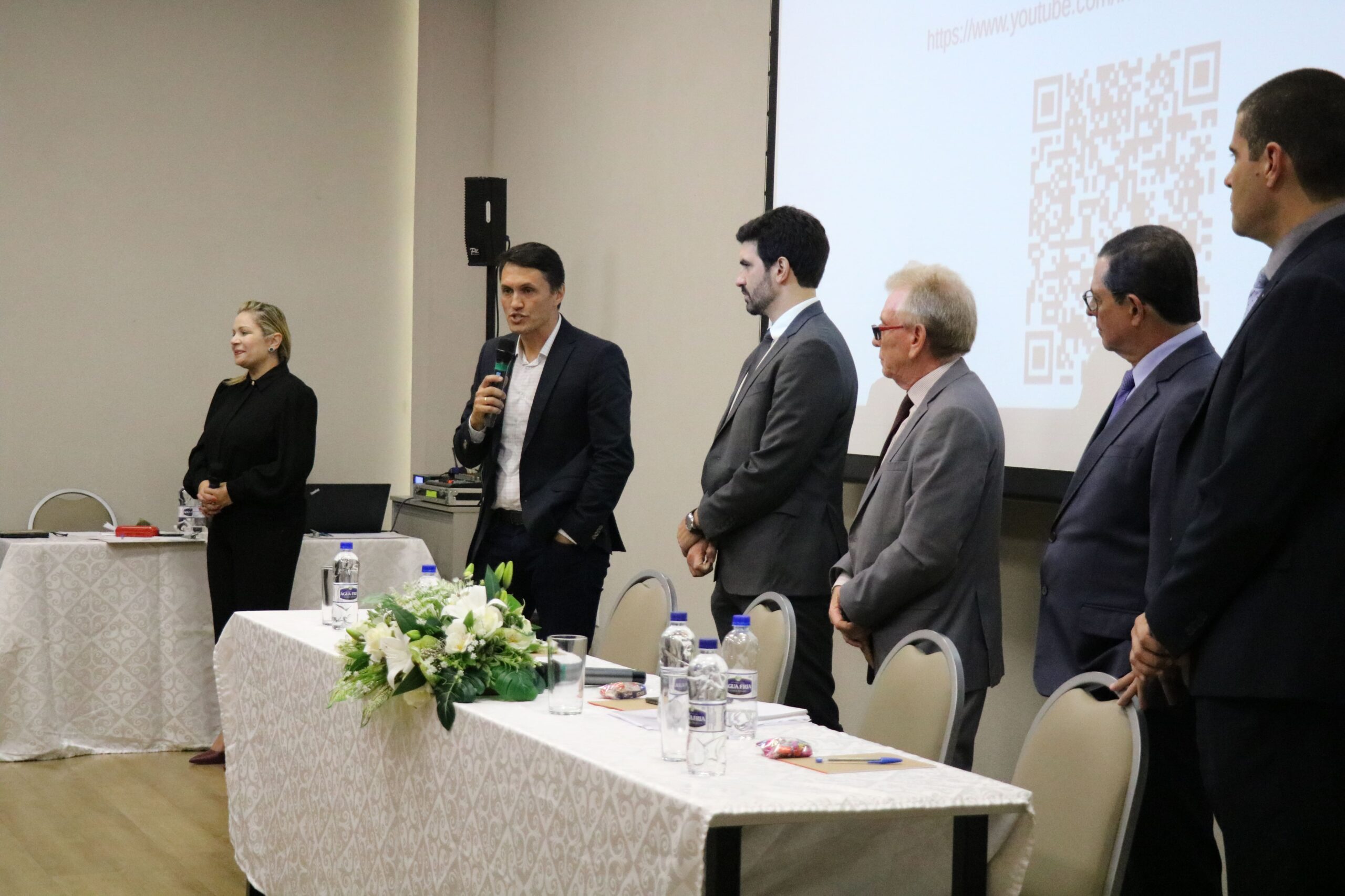 A foto mostra o presidente da Ammvet ao microfone dando as boas-vindas aos profissionais de Mogi Mirim durante o CRMV-SP Escuta. Ele está postado atrás de uma mesa com toalha branca e um arranjo de flores, ao lado do assessor jurídico, do presidente, do secretário-geral e do diretor técnico do CRMV-SP.