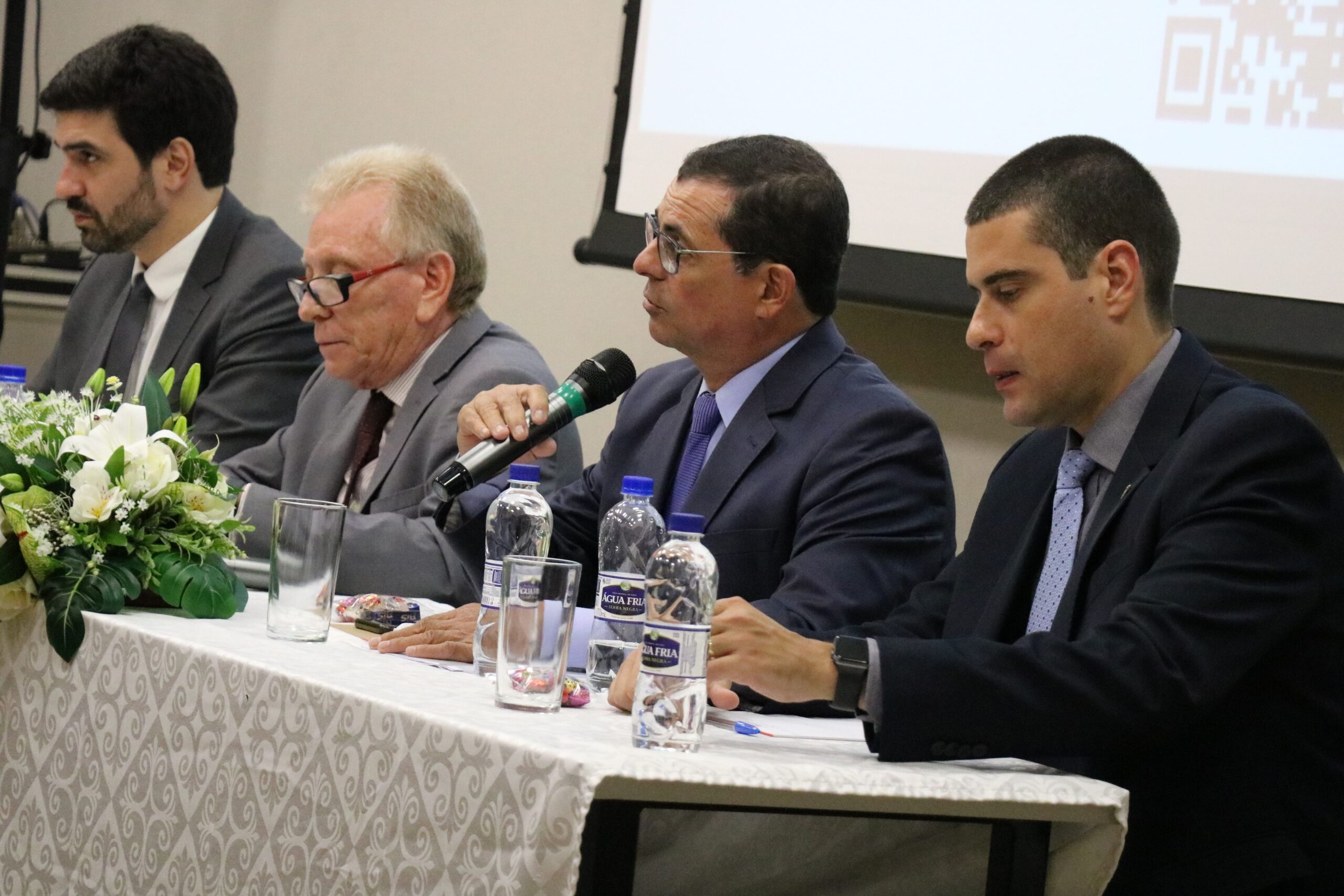 A imagem mostra o assessor, o presidente, o secretário-geral e o diretor técnico do CRMV-SP, sentados atrás de uma mesa com toalha branca e um arranjo de flores. No momento da foto, o secretário-geral é quem fala ao microfone.