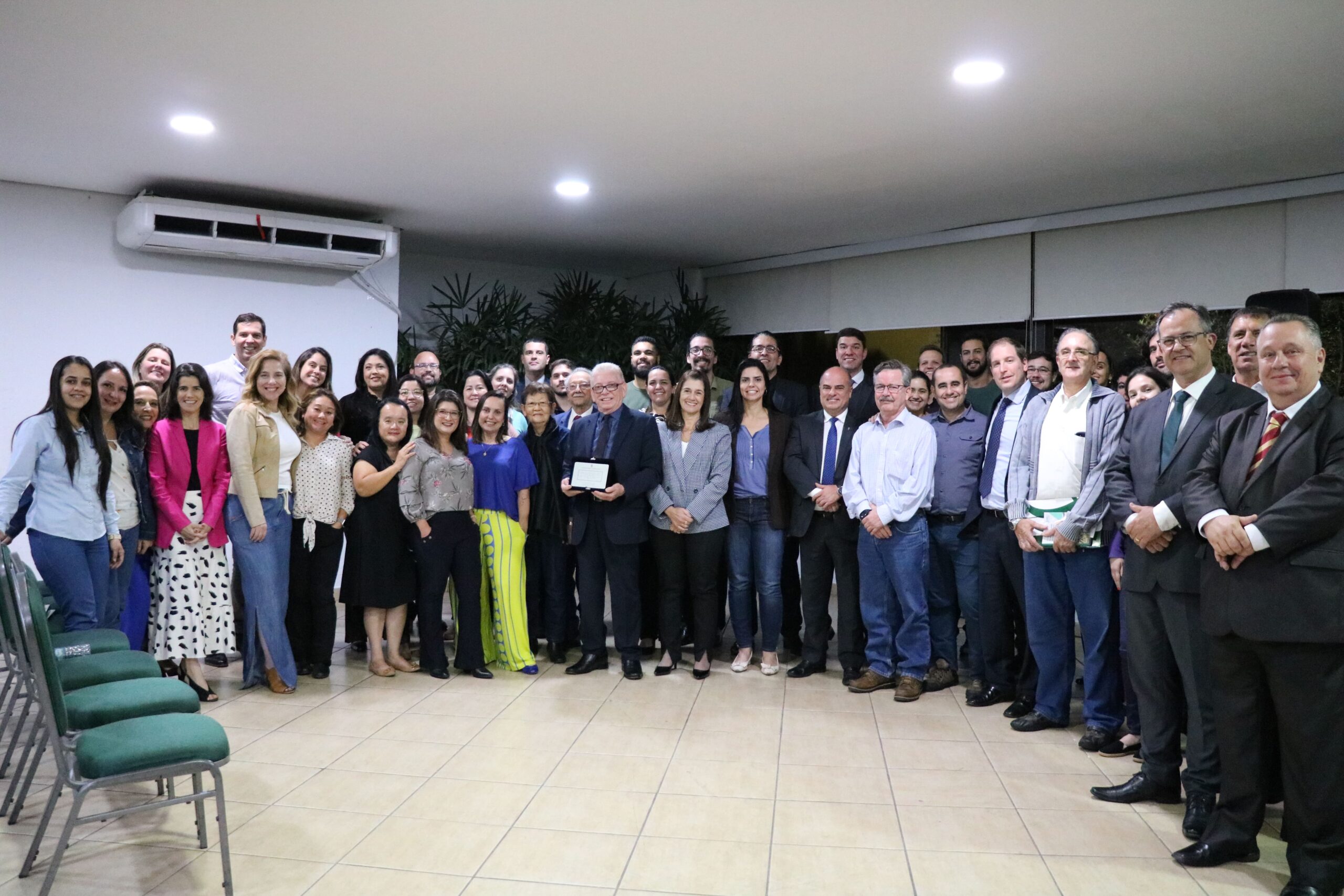 Na foto há um grupo de pessoas, em pé, posando para a foto. Dr. Odemilson Mossero está com uma placa em mãos, como homenagem