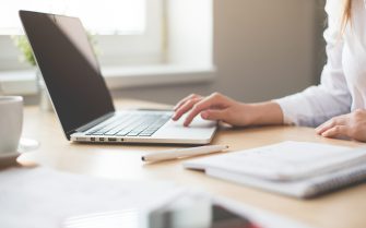 Na imagem aparece uma mesa de trabalho com papéis, celular e um computador que está sendo manuseado por uma mulher de roupa branca, que aparece apenas parcialmente.