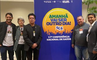 Na imagem há três pessoas, entre elas apenas uma mulher. Eles posam ao lado de um painel da 17ª Conferência Nacional de Saúde, com os dizeres 