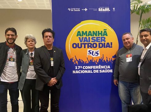 Na imagem há três pessoas, entre elas apenas uma mulher. Eles posam ao lado de um painel da 17ª Conferência Nacional de Saúde, com os dizeres 