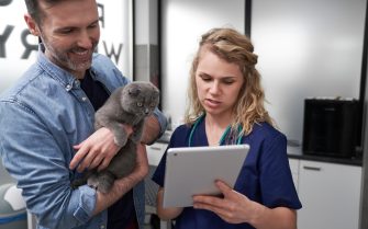 A foto mostra uma médica-veterinária segurando um tablet. Ao lado dela há um homem com um gato no colo.