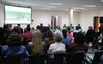 Na imagem aparece um auditório lotado de profissionais. Eles estão de costas para a câmera, sentados em cadeiras e dispostos de frente para a diretoria do Conselho, que está a frente sentada atrás de uma mesa com toalha branca e arranjo de flores. Na sala há ainda um telão em que se destaca o site do CRMV-SP com banner sobre a campanha de recadastramento.